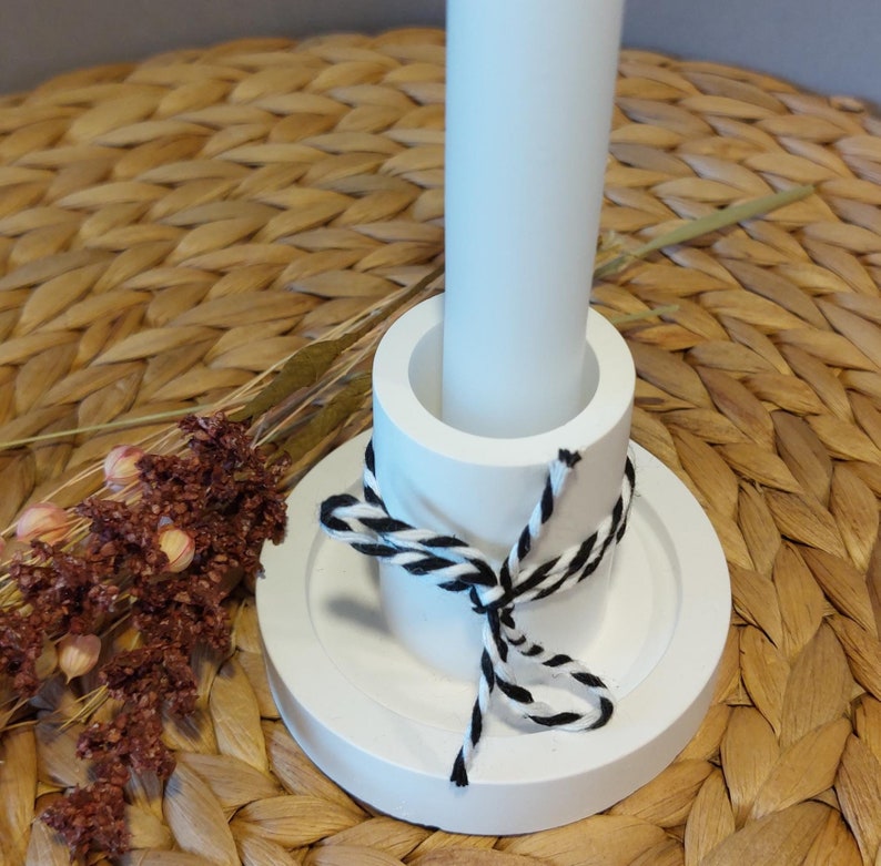 Könnte beinhalten: Ein weißer Kerzenhalter mit rundem Sockel und zylindrischer Mitte, gebunden mit schwarz-weißem Garn. Eine weiße Kerze ist in der Mitte eingesetzt. Getrocknete Blumen sind an der Seite.