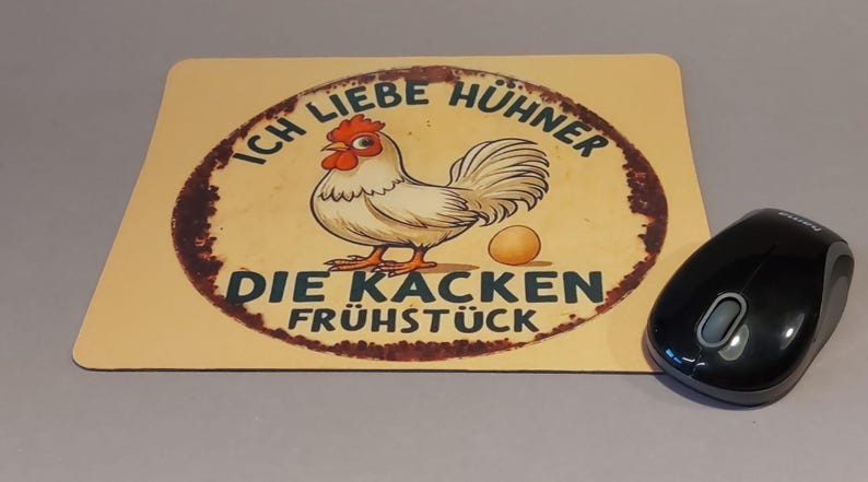 Könnte beinhalten: Rechteckiges Mauspad mit einer Cartoon-Hühner-Illustration und deutschem Text. Das Huhn ist weiß mit rotem Kamm und Kehllappen, und ein Ei liegt daneben. Eine schwarze kabellose Maus befindet sich rechts.