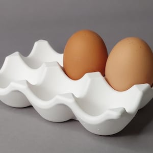 Könnte beinhalten: Ein weißer Eierhalter aus Keramik mit wellenförmigem Design, der zwei braune Eier hält. Das Eiertablett hat sechs Fächer und steht auf einer grauen Oberfläche. Die Eier sind oval geformt und haben eine glatte Textur.