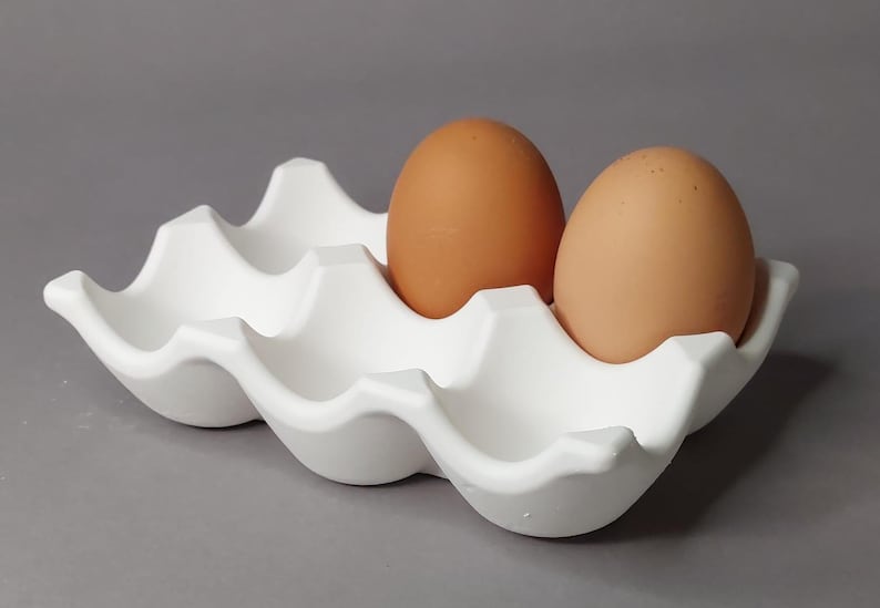 Könnte beinhalten: Ein weißer Eierhalter aus Keramik mit wellenförmigem Design, der zwei braune Eier hält. Das Eiertablett hat sechs Fächer und steht auf einer grauen Oberfläche. Die Eier sind oval geformt und haben eine glatte Textur.