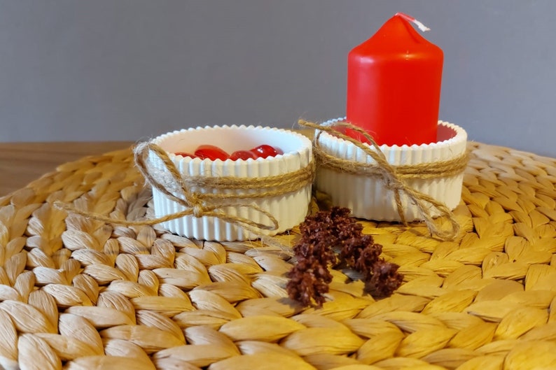 Könnte beinhalten: Zwei weiße Kerzenhalter aus Keramik mit rustikalem Garn-Akzent. Ein Halter hat rote Dekorperlen im Inneren. Eine rote Stumpenkerze sitzt im anderen Halter.