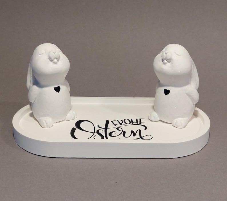 Könnte beinhalten: Zwei weiße Hasenfiguren mit schwarzen Herzakzenten stehen auf einem weißen ovalen Tablett. Auf dem Tablett steht in schwarzer Schrift "FROHE Ostern". Die Hasen blicken sich an und bilden eine bezaubernde Osterdekoration.