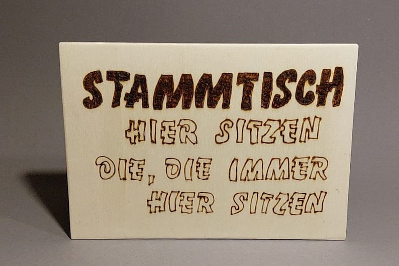 Könnte beinhalten: Ein Holzschild mit dem Text "Stammtisch Hier Sitzen Die, Die Immer Hier Sitzen", der in die Oberfläche gebrannt ist.
