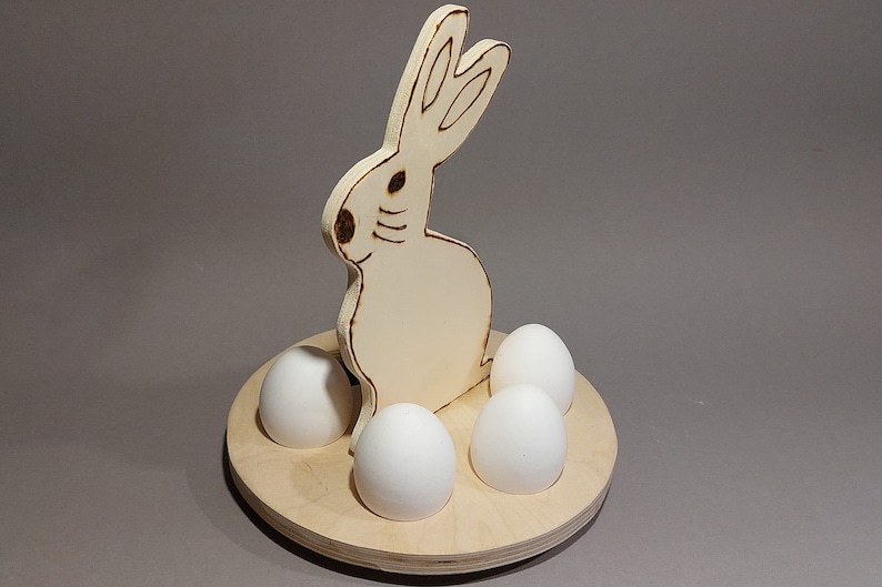 Könnte beinhalten: Osterei-Halter aus Holz mit vier weißen Eiern. Der Hase ist hell gefärbt mit dunkelbraunen Konturen. Die Eier sind um die Basis des Hasen auf einer runden Holzplattform angeordnet. Ein dekorativer Artikel für Ostern.