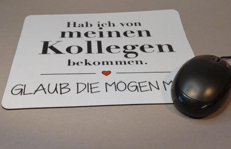 Könnte beinhalten: Ein weißes Mauspad mit schwarzem Text, der "Hab ich von meinen Kollegen bekommen.  GLAUB DIE MÖGEN MICH." lautet.  Ein rotes Herzsymbol befindet sich zwischen den beiden Textzeilen.  Eine schwarze Computermaus liegt rechts auf dem Mauspad.