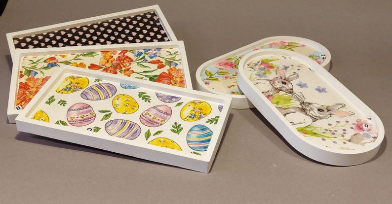 Könnte beinhalten: Eine Sammlung dekorativer Tabletts mit weißen Rahmen und gemusterten Innenräumen. Designs umfassen Blumen, Ostereier und Hasen. Die Tabletts sind rechteckig und oval, geeignet für Heimdekoration oder zum Servieren.