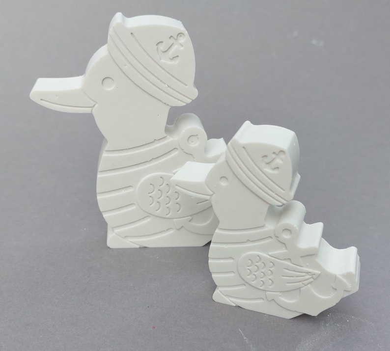 Könnte beinhalten: Zwei weiße Entenfiguren, jede mit einem Matrosenhut mit Ankeremblem. Die Enten haben detaillierte Flügel- und Körperdesigns und sind vor einem neutralen grauen Hintergrund platziert. Die Figuren bestehen aus einem festen, glatten Material.