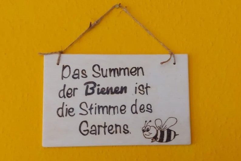 Könnte beinhalten: Ein weißes Holzschild mit schwarzem Text, der "Das Summen der Bienen ist die Stimme des Gartens." lautet. Im unteren rechten Eck des Schildes ist eine schwarz-weiße Cartoon-Biene gezeichnet.