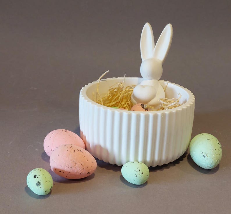 Könnte beinhalten: Eine weiße Keramik-Hasenfigur sitzt in einer gerippten weißen Schale, die mit Stroh und gesprenkelten Eiern gefüllt ist. Die Schale ist von zusätzlichen gesprenkelten Eiern in Rosa- und Grüntönen umgeben, was eine festliche Osterdekoration ergibt.