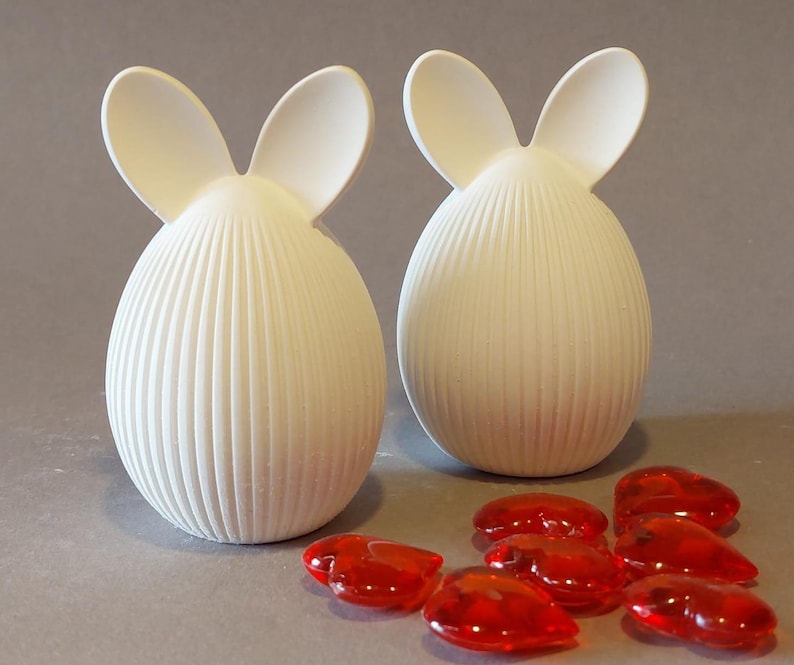 Könnte beinhalten: Zwei weiße, eiförmige Deko-Objekte mit hasenohrförmigen Aufsätzen. Die Körper haben vertikale geriffelte Details. Mehrere rote, herzförmige Glassteine liegen vor den Objekten.