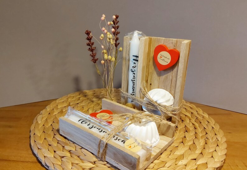 Könnte beinhalten: Ein Holz-Geschenkset mit einer weißen Kerze, zwei weißen Keramikschalen und einem roten Herzen. Die Kerze trägt den Text "Herzgesummse". Das Geschenkset ist mit Schnur umwickelt und mit getrockneten Blumen verziert.