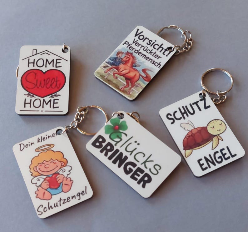 Könnte beinhalten: Eine Sammlung rechteckiger Schlüsselanhänger mit verschiedenen Designs und Texten. Ein Schlüsselanhänger zeigt ein Haus und ein Herz mit "HOME Sweet HOME". Andere Schlüsselanhänger haben Bilder von Pferden, Engeln, Schildkröten und einem vierblättrigen Kleeblatt. Jeder Schlüsselanhänger hat einen Metallring.