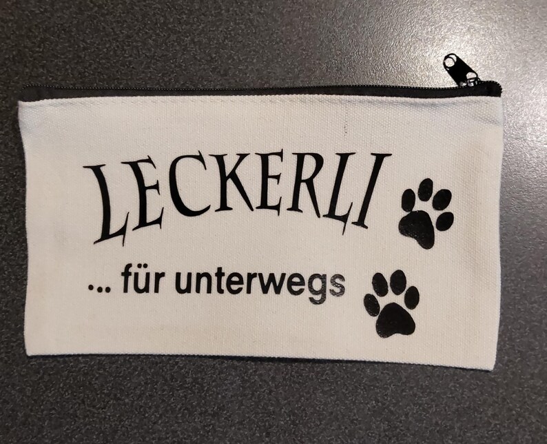 Könnte beinhalten: Weiße Canvas-Reißverschlusstasche mit schwarzem Text "LECKERLI... für unterwegs" und zwei schwarzen Pfotenabdrücken.