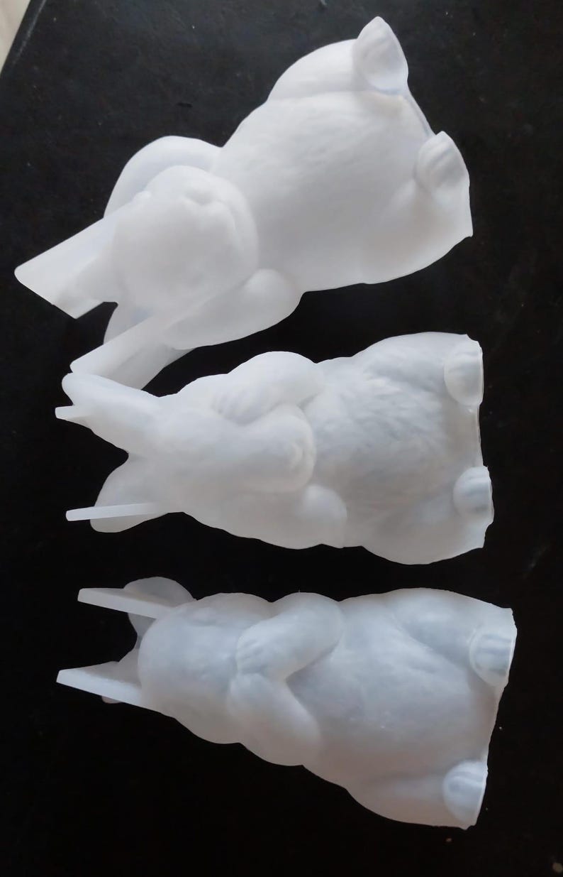 Könnte beinhalten: Drei weiße Hasenfiguren in verschiedenen Posen. Die Hasen liegen, wobei ihre Ohren und Pfoten sichtbar sind. Die Figuren bestehen aus einem glatten, matten Material und sind vor einem dunklen Hintergrund platziert.