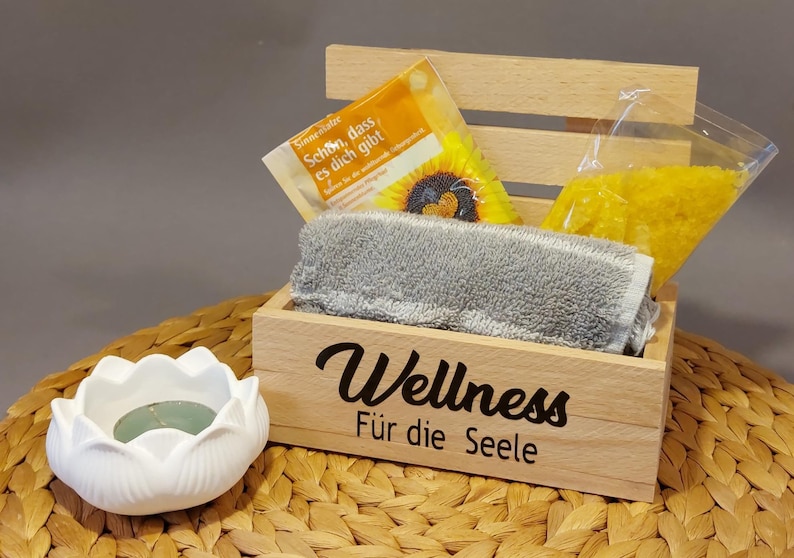 Könnte beinhalten: Holzkiste mit dem Text "Wellness Für die Seele" auf der Vorderseite. Die Kiste enthält ein graues Handtuch, eine Tüte gelber Badesalze und eine kleine Packung Badesalze mit Sonnenblumenmuster. Die Kiste steht auf einer geflochtenen Weidenoberfläche neben einem weißen Kerzenhalter aus Keramik in Lotusform.