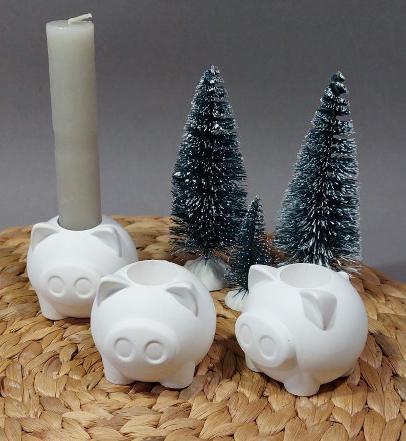 Könnte beinhalten: Drei weiße Kerzenhalter aus Keramik in Form von Schweinen mit einer einzelnen weißen Kerze im mittleren Halter. Die Kerzenhalter stehen auf einer geflochtenen Weidenoberfläche mit zwei kleinen künstlichen Kiefern dahinter.