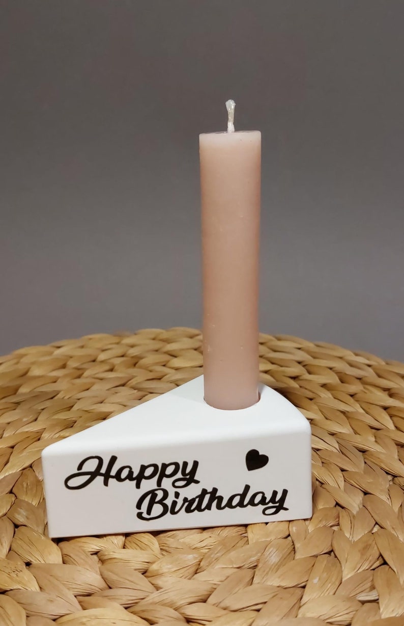 Könnte beinhalten: Weißer Kerzenhalter aus Keramik in Form eines Kuchenstücks mit dem Text "Happy Birthday" und einem kleinen Herz. Eine einzelne, hellbraune Kerze befindet sich im Halter.