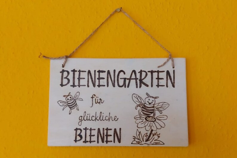 Könnte beinhalten: Ein weißes Holzschild mit schwarzem Text, der "Bienengarten für glückliche Bienen" lautet. Das Schild hat zwei handgezeichnete Bienen darauf, eine fliegt und eine sitzt auf einer Blume.