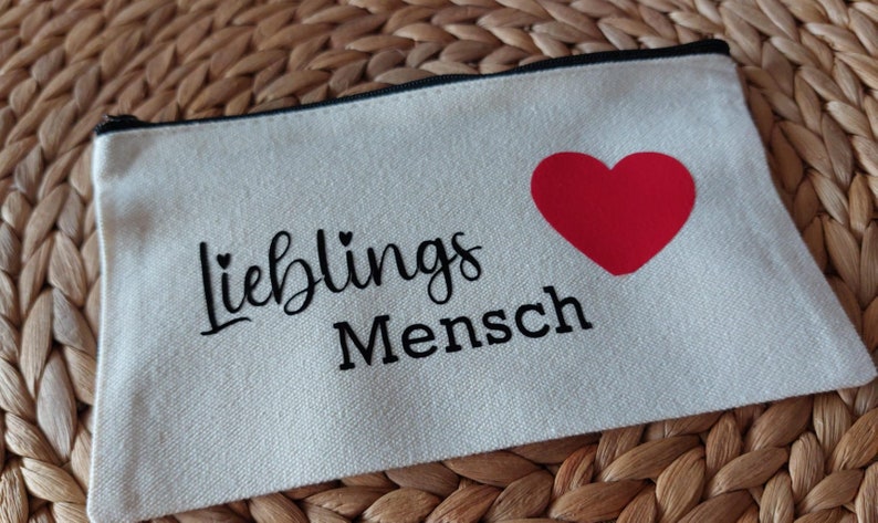Könnte beinhalten: Eine weiße Canvas-Reißverschlusstasche mit dem Text "Lieblings Mensch" in schwarzer Schrift und einem roten Herzen.