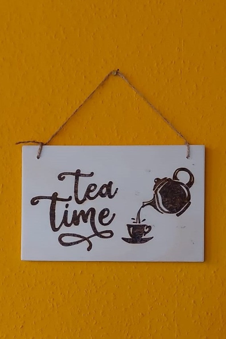 Könnte beinhalten: Ein weißes Holzschild mit dem Text "Tea Time" in schwarzer Kursivschrift. Rechts auf dem Schild ist eine schwarze Silhouette einer Teekanne, die Tee in eine Tasse gießt.