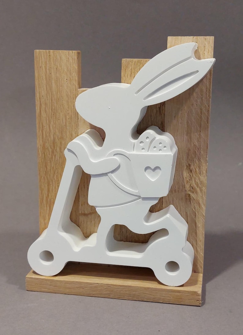 Könnte beinhalten: Eine weiße Holzfigur eines Osterhasen auf einem Roller, der einen Korb mit Eiern trägt. Der Hase ist auf einem Holzblock montiert. Der Korb hat eine Herzform. Der Hintergrund ist hellgrau.