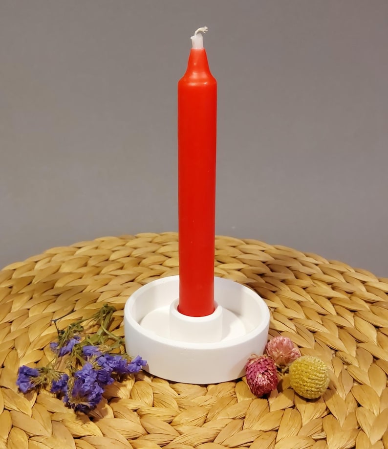 Könnte beinhalten: Ein weißer Kerzenhalter aus Keramik mit einer roten Kerze. Der Kerzenhalter steht auf einer geflochtenen Weidenoberfläche.