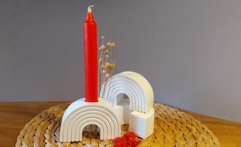 Könnte beinhalten: Weißer Kerzenhalter aus Keramik in Regenbogenform mit einer roten Kerze in der Mitte. Der Halter steht auf einem geflochtenen Untersetzer.