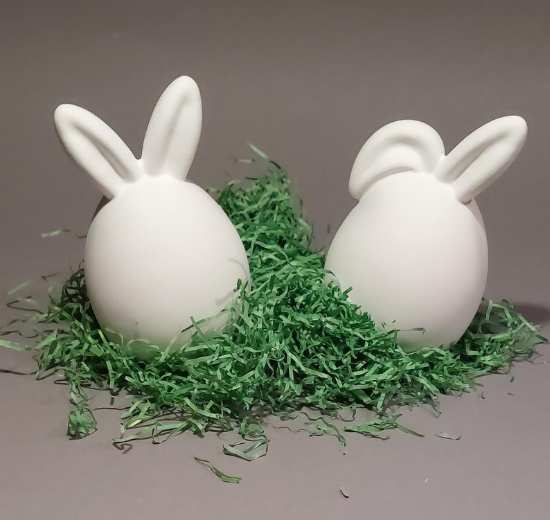 Könnte beinhalten: Zwei weiße Keramik-Ostereier-Figuren mit Hasenohren, eingebettet in grünes Papiergras, vor grauem Hintergrund. Die eiförmigen Figuren haben eine glatte Oberfläche, ideal für Osterdekoration oder Bastelarbeiten.