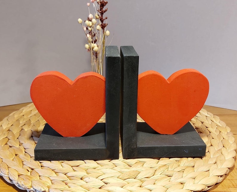 Könnte beinhalten: Ein Paar Buchstützen mit einem leuchtend orangefarbenen Herzdesign. Die herzförmigen Akzente sind auf einer schwarzen rechteckigen Basis angeordnet. Dekorative Trockenblumen befinden sich im Hintergrund. Die Buchstützen sind aus Holz.