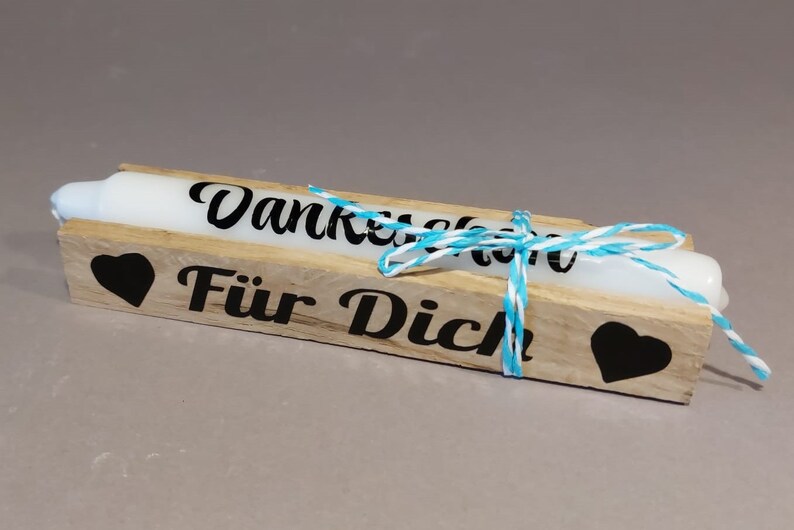 Könnte beinhalten: Zwei weiße Kerzen in einer Holzkiste mit einer blauen und weißen Kordelschleife. Die Kiste ist mit schwarzen Herzen und dem Text "Danke schön für dich" verziert.