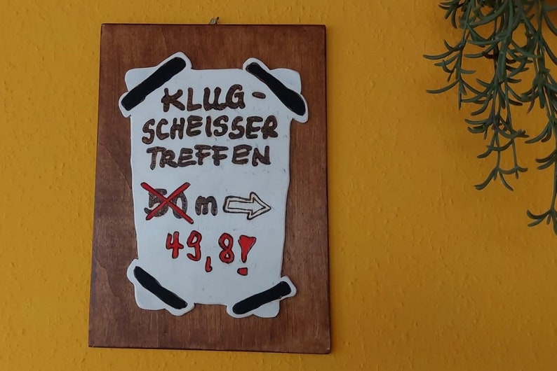 Könnte beinhalten: Ein Holzschild mit weißem Papier daran befestigt. Das Papier ist mit schwarzem Stift beschriftet und lautet "KLUG-SCHEISSER TREFFEN" und "50m" mit einem Pfeil nach rechts. Außerdem sind rote Zahlen zu sehen, die "43,8!" anzeigen.