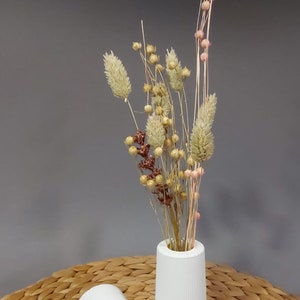 Könnte beinhalten: Zwei weiße Keramikvasen mit gerippter Textur. Die Vase rechts enthält getrocknete Blumen mit rosa, braunen und beigefarbenen Akzenten.