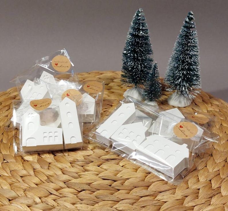 Könnte beinhalten: Weiße Holzhäuser mit einem Herzen und dem Schriftzug "Handmade with Love" auf einem Etikett. Die Häuser sind in durchsichtigen Plastiktüten verpackt und liegen auf einer geflochtenen Weidenoberfläche. Im Hintergrund stehen zwei kleine weiße, gefrostete künstliche Weihnachtsbäume.