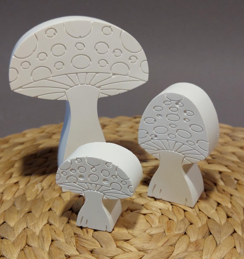 Könnte beinhalten: Drei weiße Holzpilzfiguren mit einem gepunkteten Muster auf der Oberseite. Die Pilze sind unterschiedlich groß und in einer Reihe auf einer gewebten Oberfläche angeordnet.