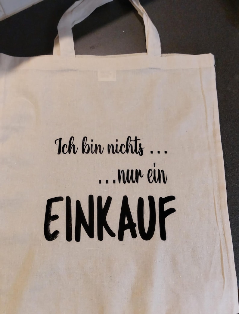 Könnte beinhalten: Eine weiße Stofftasche mit schwarzem Text, der "Ich bin nichts... ...nur ein EINKAUF" lautet.