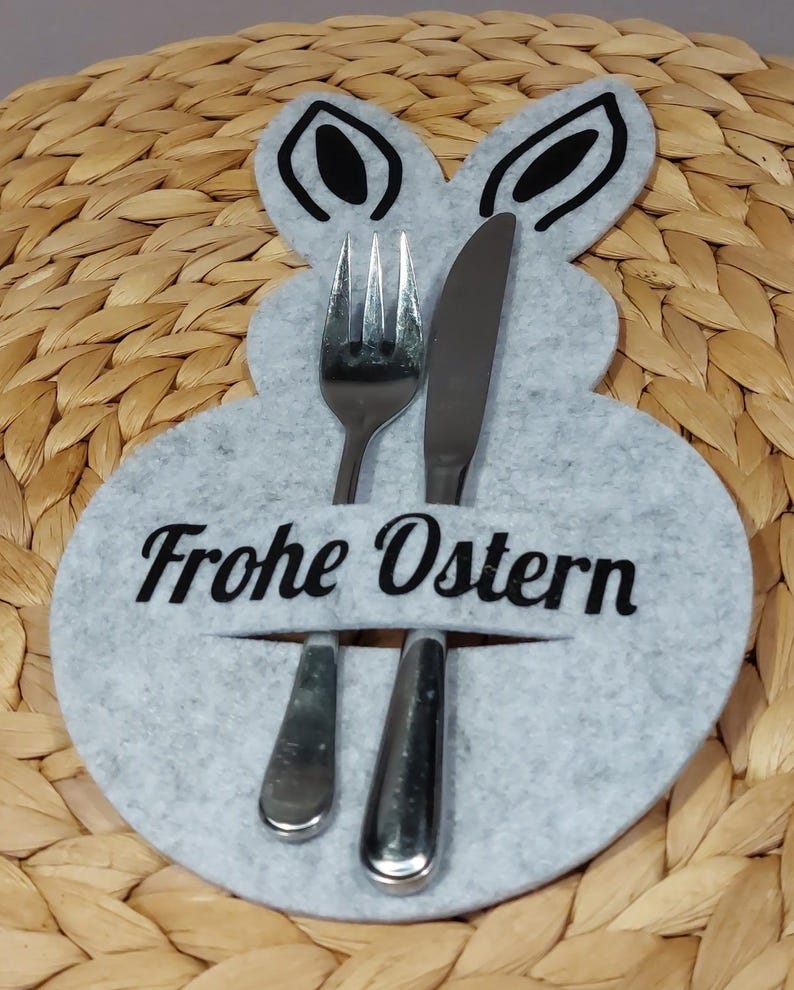 Könnte beinhalten: Ein grauer Oster-Besteckhalter aus Filz in Hasenform. Der Halter hat eine Gabel, ein Messer und einen Löffel. Der Text "Frohe Ostern" ist in Schwarz auf dem Halter aufgedruckt.