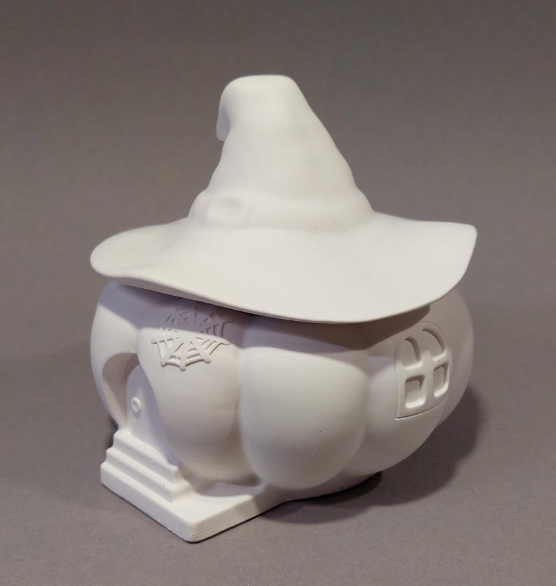 Könnte beinhalten: Ein weißer Keramikbehälter in Kürbisform mit einem Hexenhutdeckel. Der Behälter hat eine kleine Tür, ein Fenster und ein Spinnennetzdesign. Das Design ist skurril und für Halloween geeignet.