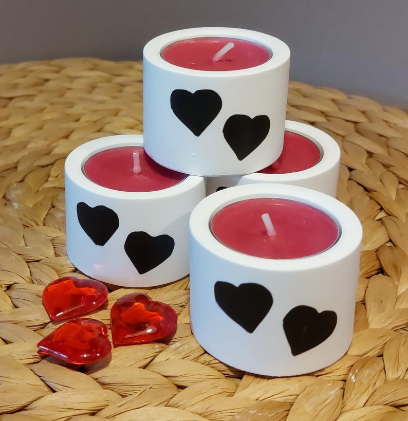 Könnte beinhalten: Vier weiße Kerzenhalter mit schwarzen Herzmotiven. Jeder Halter hat eine rote Teelichtkerze drin. Im Vordergrund liegen drei rote herzförmige Glasperlen.