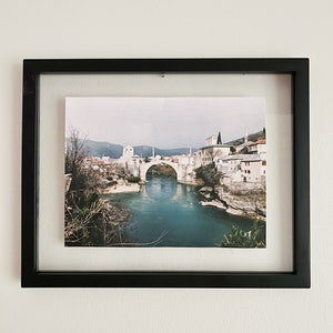 May include: A framed photograph of a bridge over a river in a city. The bridge is arched and made of stone. The river is blue and the city is in the background.