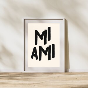 Peut inclure: Une impression encadr&eacute;e blanche avec le texte "MI AMI" en lettres noires en forme de coups de pinceau sur un fond cr&egrave;me.