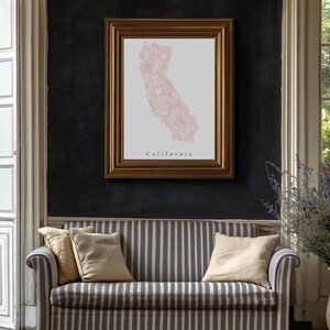 May include: A framed print of California in a gold frame hangs above a striped sofa. The print features a pink floral design within the state's outline, with the word "California" below. The sofa has beige and blue vertical stripes and several beige pillows.