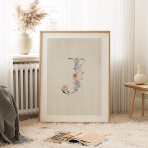 May include: A framed print with a watercolor illustration of the letter J surrounded by pink and blue flowers. The background is a light beige color.