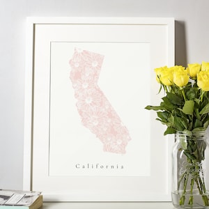May include: A framed print featuring a pink floral design in the shape of California. The word "California" is printed in a simple font below the state outline. A bouquet of yellow roses in a glass jar sits to the right.