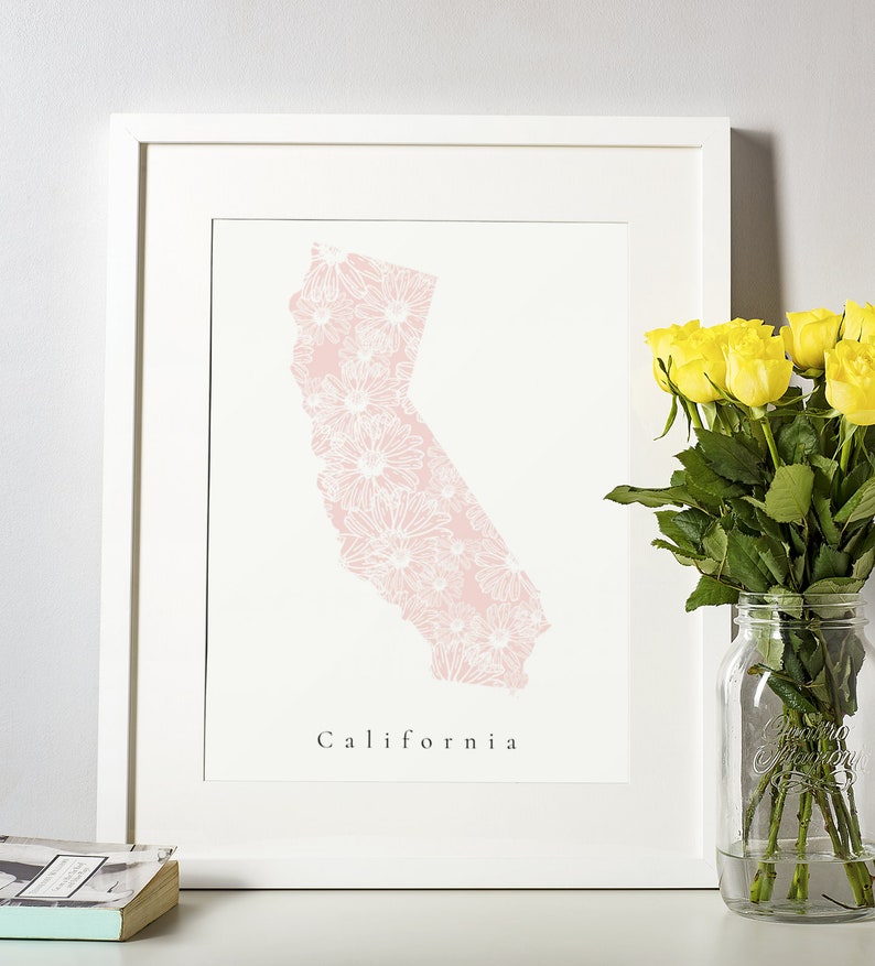 May include: A framed print featuring a pink floral design in the shape of California. The word "California" is printed in a simple font below the state outline. A bouquet of yellow roses in a glass jar sits to the right.