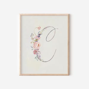 May include: A framed watercolor print featuring the letter "C" adorned with a floral arrangement. The flowers include pink roses, purple accents, and green foliage. The artwork is set against a soft, off-white background, and framed in a light wood.