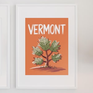 May include: A framed art print featuring the word "VERMONT" in white brush lettering above a stylized tree illustration. The tree has green leaves and a brown trunk against an orange background.