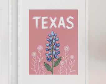 Puede incluir: Una impresión enmarcada con una ilustración de lupino azul y la palabra "TEXAS" en trazos de pincel blancos sobre un fondo rosa. La obra de arte incluye acentos florales blancos y un tallo verde.
