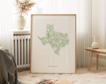 Puede incluir: Un estampado enmarcado con el contorno de Texas, relleno con un diseño floral en verde y blanco. La palabra "Texas" está impresa debajo. La obra de arte se presenta en un marco de madera clara, ideal para la decoración del hogar.