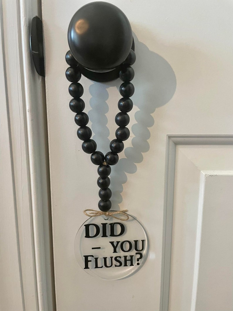 May include: A black beaded garland hangs from a black door knob with a clear round sign that reads "Did - You Flush?"