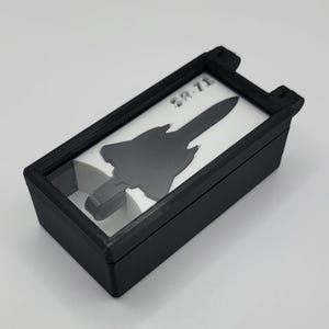 Puede incluir: Una caja rectangular negra con tapa transparente, con una silueta negra de SR-71 sobre un fondo blanco. El texto "SR-71" es visible en la parte superior. La caja es probablemente una vitrina o un contenedor de almacenamiento.
